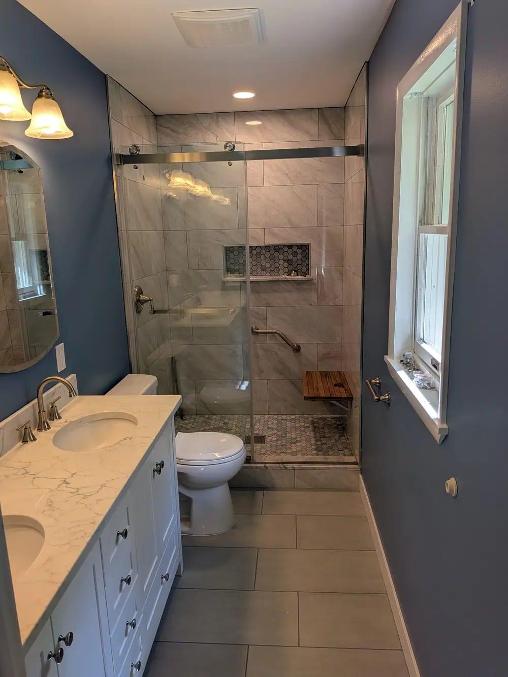 A remodeled bathroom with a tiled walk-in shower, glass slider, wood bench and double vanity